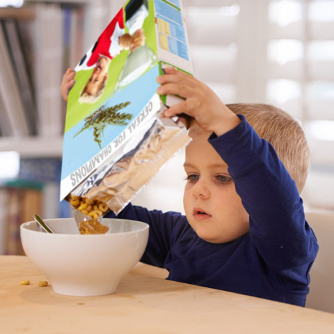 A boy pouring cereal from a rectangular box into a bowl.